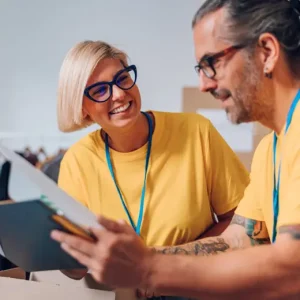 Two people wearing yellow shirts and blue lanyards smile as they review Netsuite documents or a clipboard together, appearing to work collaboratively in an indoor setting.