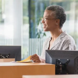 A smiling woman with short gray hair and glasses sits at a desk in an office, holding papers. Two computer monitors display NetSuite dashboards, while large windows in the background let in natural light.
